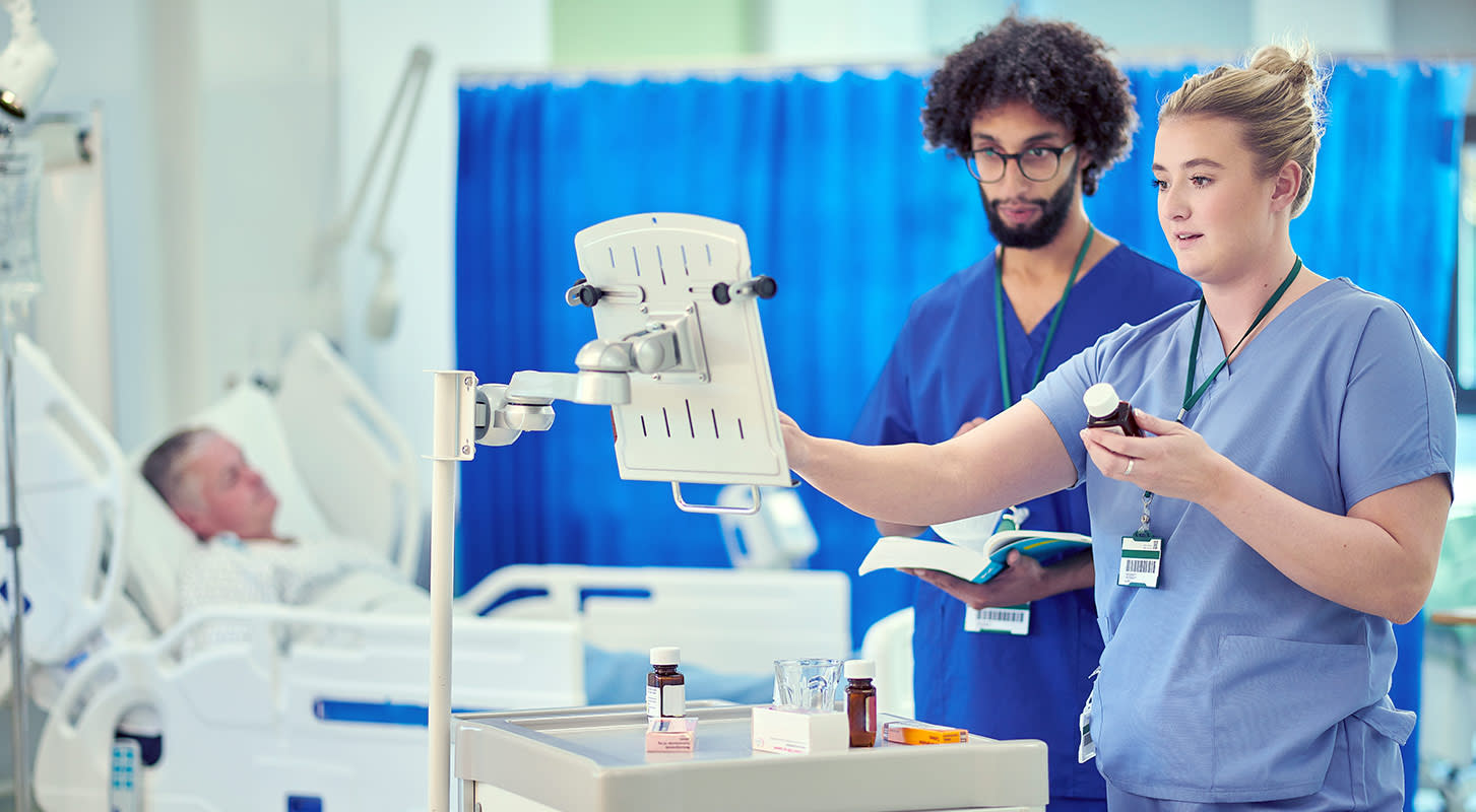 Two nurses in scrubs confirm prescriptions on a computer for a patient who is lying in a hospital bed in the background.